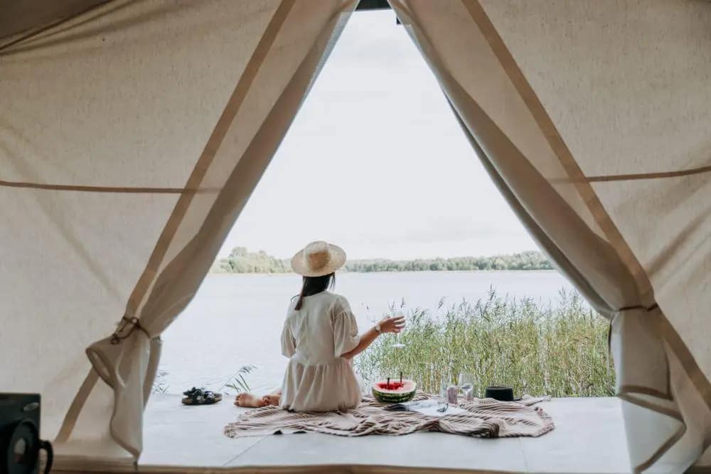 View from inside a luxury Seaside Glamping in Bali tent, showing a woman in a straw hat sitting and looking out at a serene lake or water body with reeds, with a small picnic of watermelon and a drink.