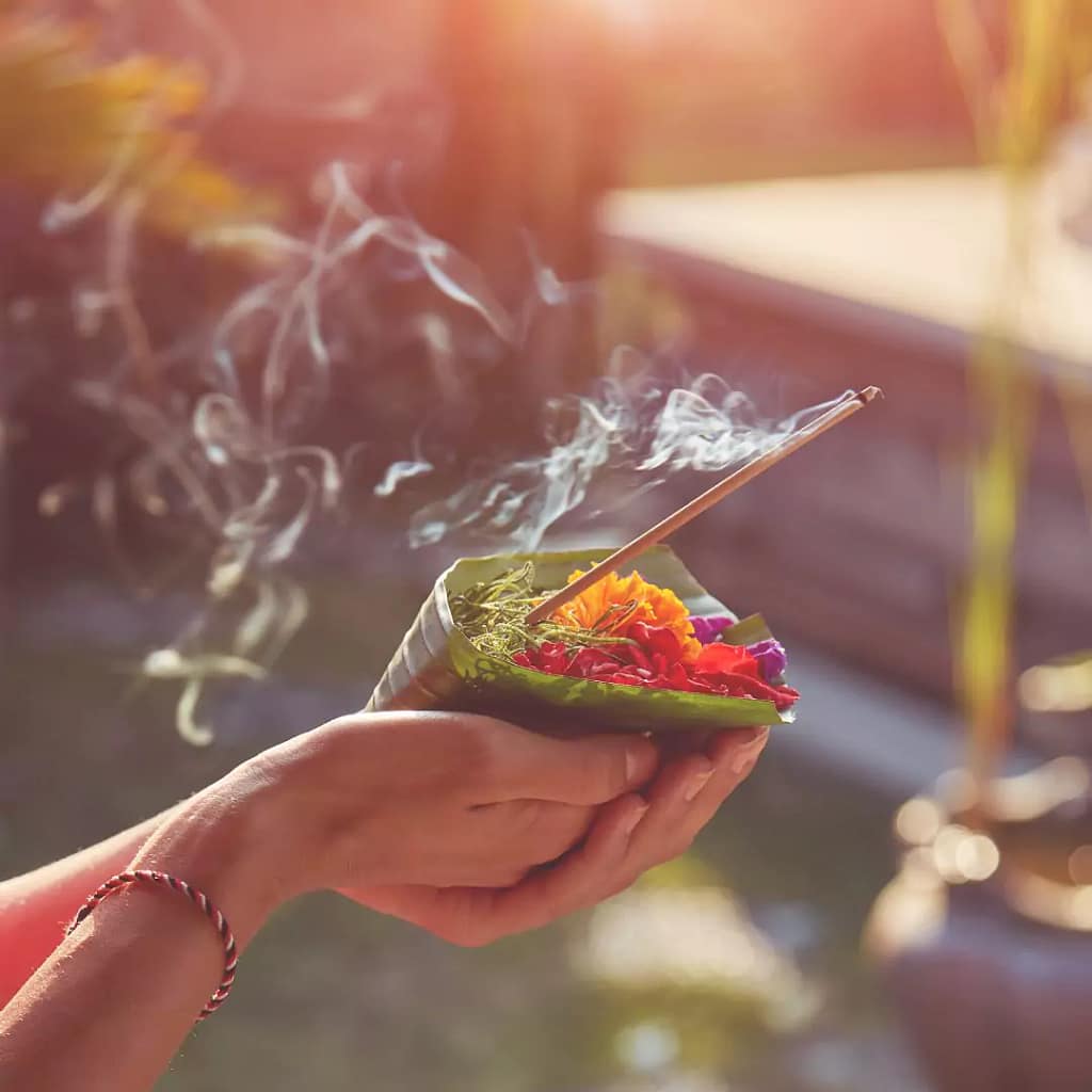 The photo of two hands carrying a canang sari with incense and smoke symbolizes an offering to the powerful. - Offerings in Bali