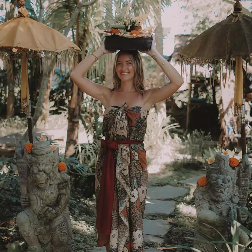 A female foreign tourist carrying a Balinese 'canang sari' offering while wearing traditional sarong, exploring cultural traditions in Bali. - religion of bali Indonesia
