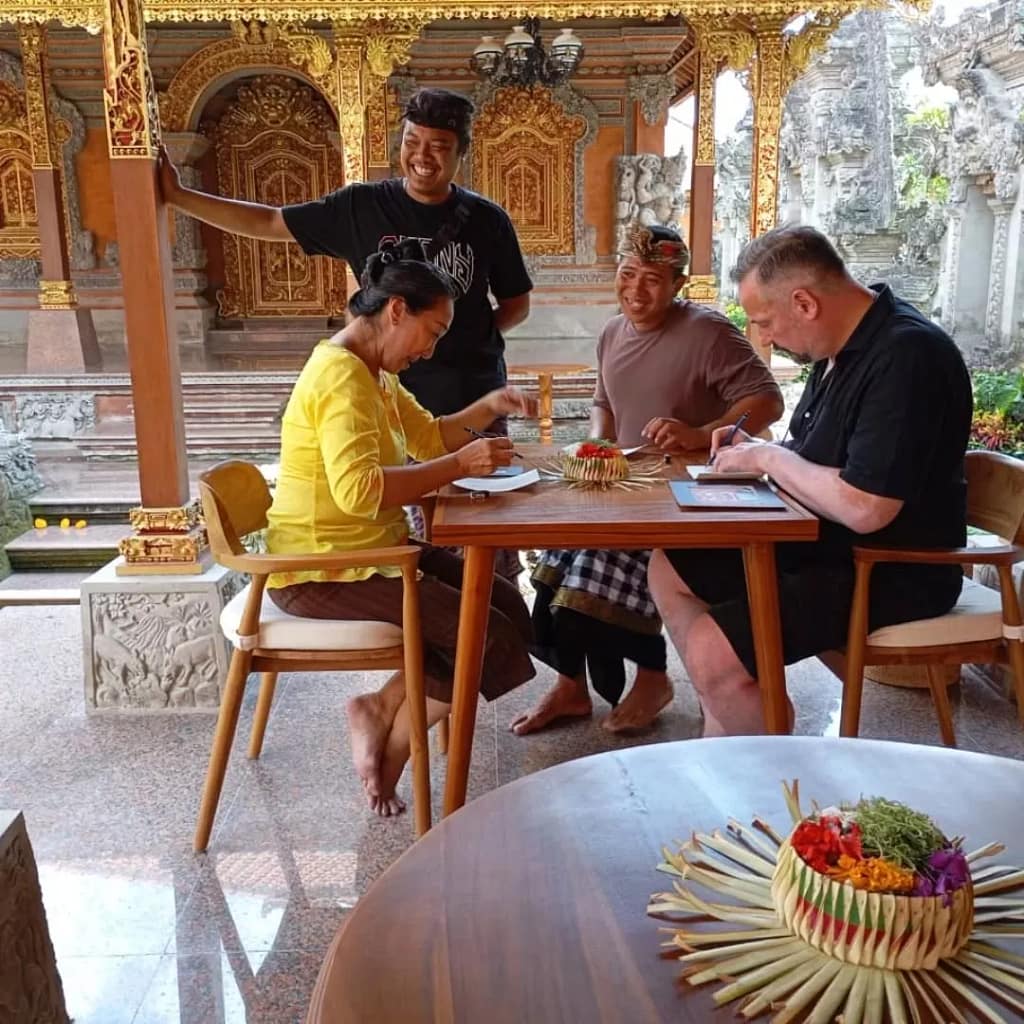 A smiling man wearing sunglasses, a white shirt, and khaki shorts, sitting cross-legged in a narrow, immersive tunnel made of countless colorful, stacked Balinese handcrafted pottery vases and pots, showcasing the vibrant artistic scene.