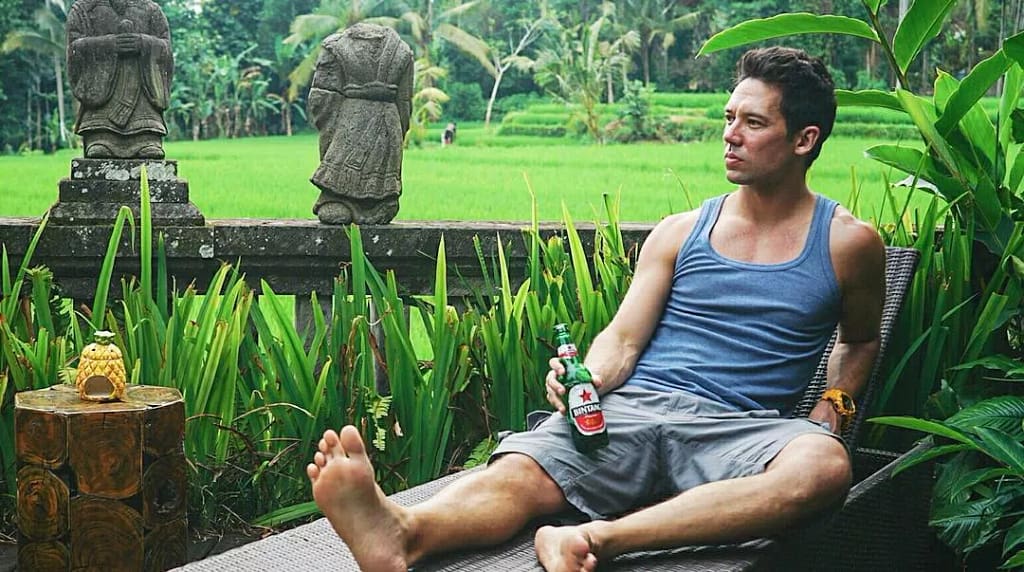 A man in a blue tank top and grey shorts is relaxing barefoot on a lounge chair, holding a bottle of Bintang beer. He gazes contemplatively over a lush green rice paddy, bordered by traditional stone statues and tropical plants, embodying the serene and relaxed atmosphere of rural Bali. Slow Living Ubud