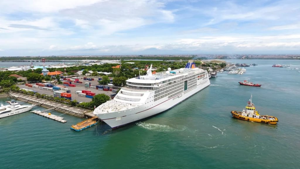 Aerial view of a large cruise ship docking at Benoa Cruise Port, with cargo containers and tugboats visible, illustrating the busy environment for Benoa Cruise Port Transfer.