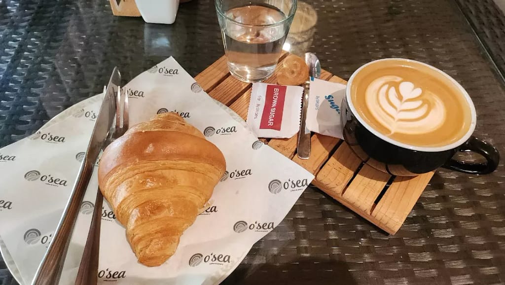 Breakfast spread at O'sea: A golden croissant served with a latte or cappuccino featuring latte art on a wooden coaster