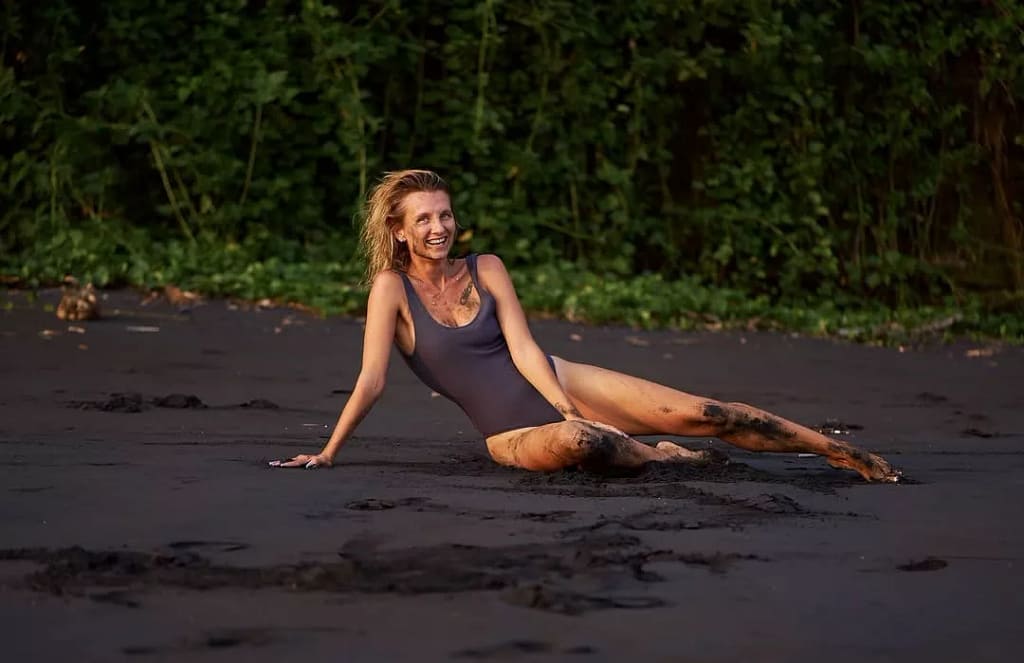 Happy woman in a dark swimsuit relaxing on Bali's black volcanic sand, covered in dirt and smiling brightly, suggesting an authentic, off-the-checklist experience away from the polished resort look. Alt: The real answer to is Bali overrated lies in abandoning the algorithmic loop for true discovery.
