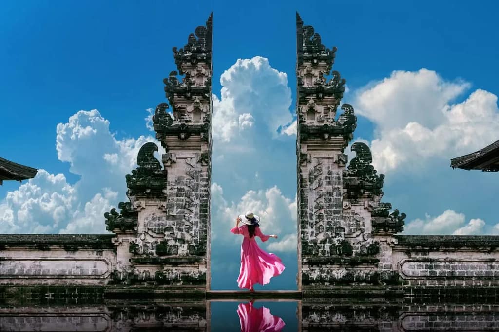 The ancient Candi Bentar gate of Bali (possibly in Lempuyang or Handara) against a bright blue sky and a large white cloud in the center. A woman in a bright pink dress and hat stands dramatically between two stone pillars, reflecting the light in front of her. Alt: Is Bali overrated? This photo depicts a picture of Bali as a perfect, staged, and impeccable 'paradise', reflecting the internet narrative discussed in the article.