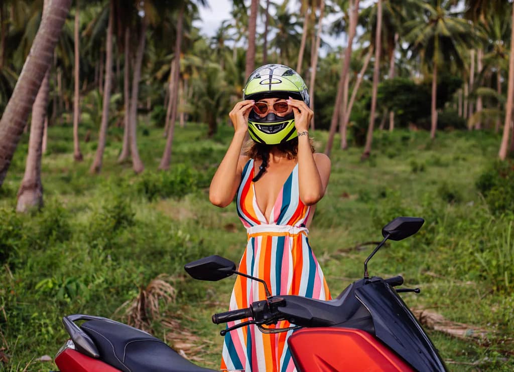 A foreign woman adjusts her lime-green helmet while standing next to a red scooter