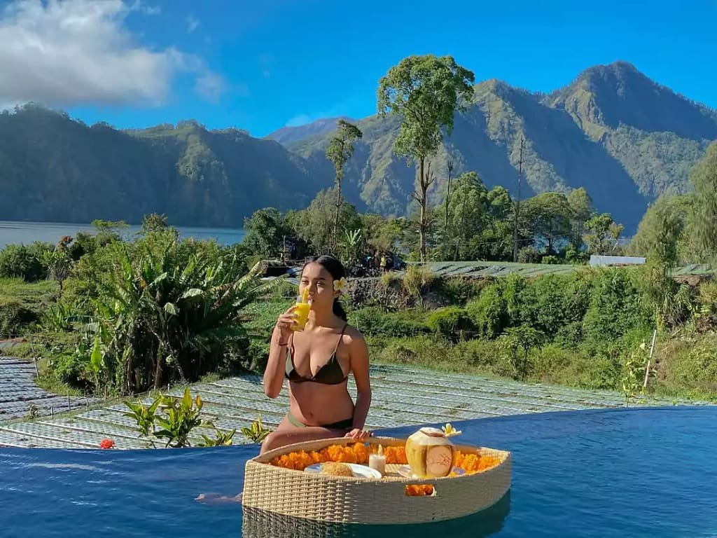 a female traveler poses by the pool of a hotel and enjoys a drink and floating breakfast with a view of the mountain and Lake Batur in Bali.