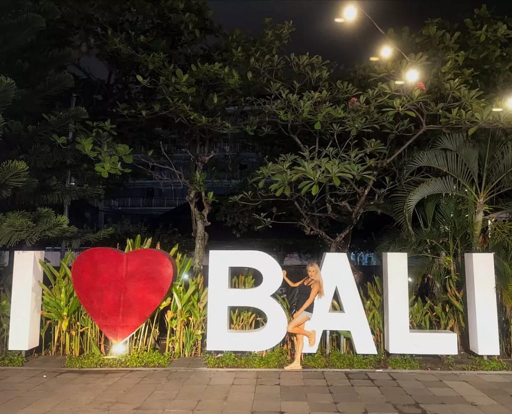 Woman posing in front of the large 'I ❤ BALI' sign at night after arriving via the port, highlighting the need for a reliable Benoa Cruise Port Transfer.