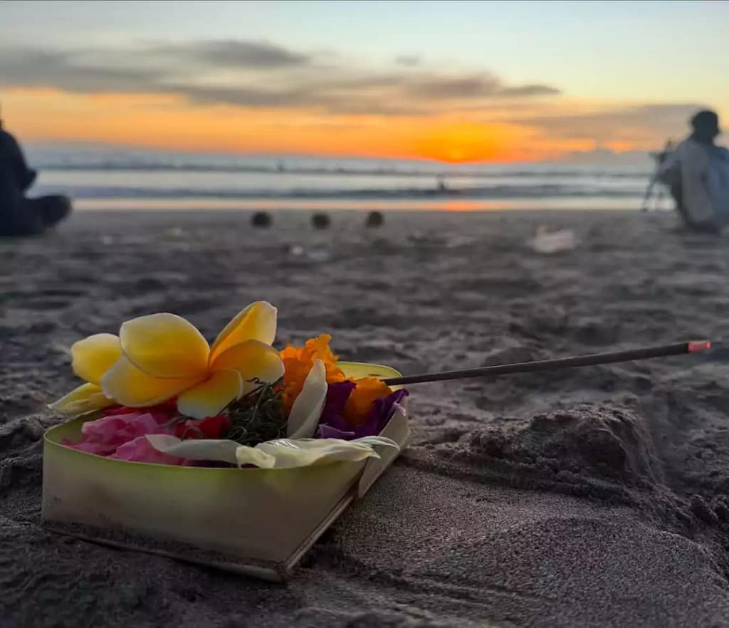 close up photo of canang sari with incense burning on the sand of kuta beach bali with beautiful sunset background. - Bali Hindu Offerings