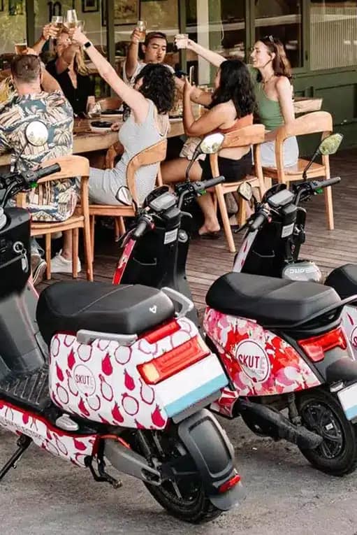 Two parked electric scooters at Bali cafe with happy tourists holding drinks - sustainable motorbike rental for eco-conscious travelers