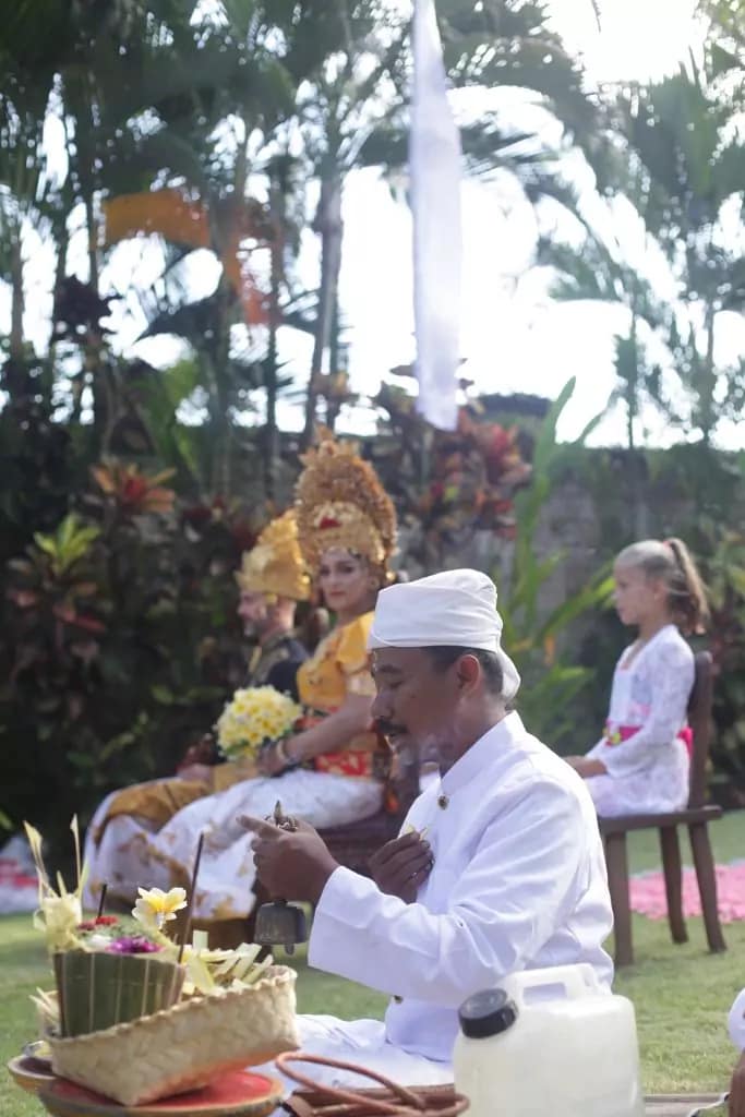 Photo of a Balinese priest praying and holding incense with white-white clothes in the background of a Caucasian groom and bride.