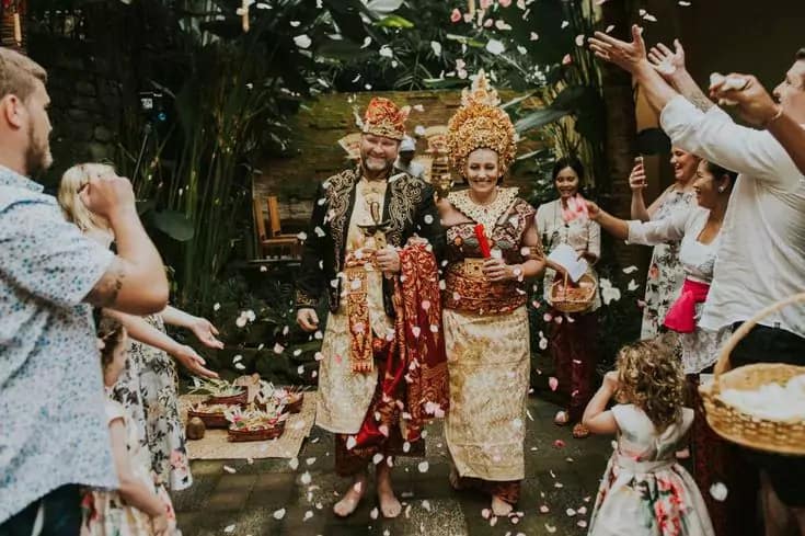 photo of the bule bride and groom wearing luxurious bali traditional clothes walking and being showered with flowers by their guests and relatives at a hotel in bali - Bali traditional wedding