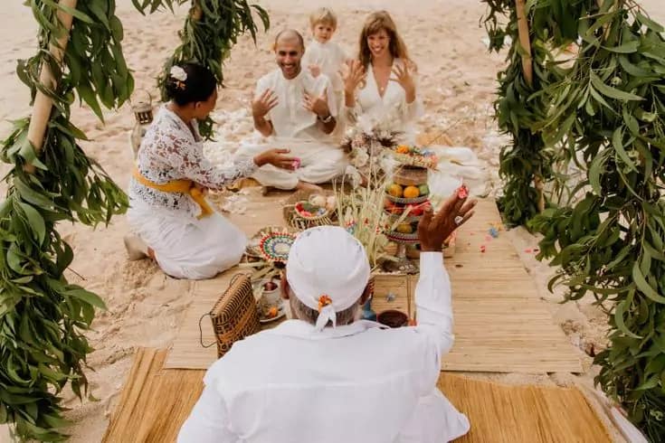 Photo of a Balinese priest in white clothes sprinkling holy water on a Caucasian bride and groom on a beach in Bali.
