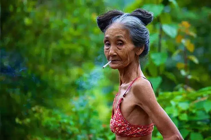 close up photo of a grandmother in Bali with a cigarette in her mouth and looking coldly at the camera and wearing only a bra