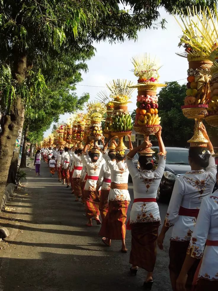 photo of a line of Balinese women carrying banten on their heads while walking down the highway to the temple. all women are wearing uniform Balinese traditional clothes.