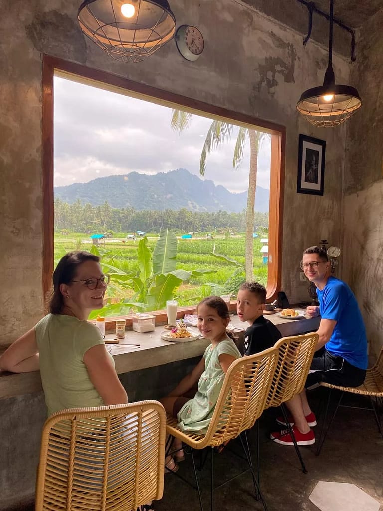 A family of four (two adults and two children) sitting at a counter inside a cafe with a large window overlooking a lush green landscape of rice fields and tropical mountains. The family is enjoying a meal. The alt text incorporates the user query: Is Ubud Worth Visiting?