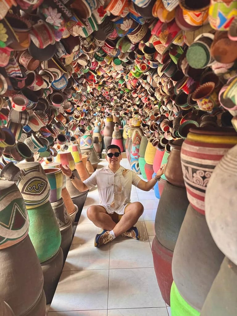 A smiling man wearing sunglasses, a white shirt, and khaki shorts, sitting cross-legged on the floor in a narrow, tunnel-like space. The space is entirely filled with countless colorful, handcrafted pottery vases and pots of various sizes and designs, stacked from floor to ceiling, creating an immersive and vibrant artistic display. Why Tourists Love Ubud Bali