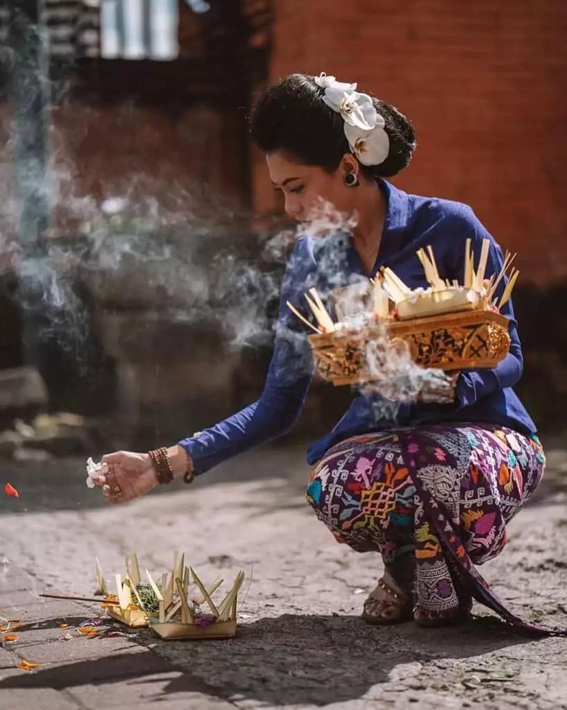 A photo of a mother in Bali placing canang sari and sprinkling holy water on the offerings by carrying a pile of canang sari in a woven basket. She is wearing full Balinese traditional blue dress and the smoke of incense is blowing in the wind.
