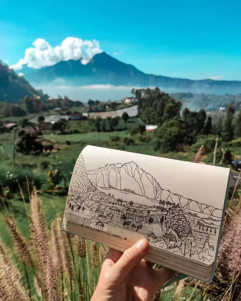 photo of a person holding a book containing beautiful panoramic landscape paintings of Lake Batur and Mount Batur with the background of the original scenery at Lake Batur in Bali.