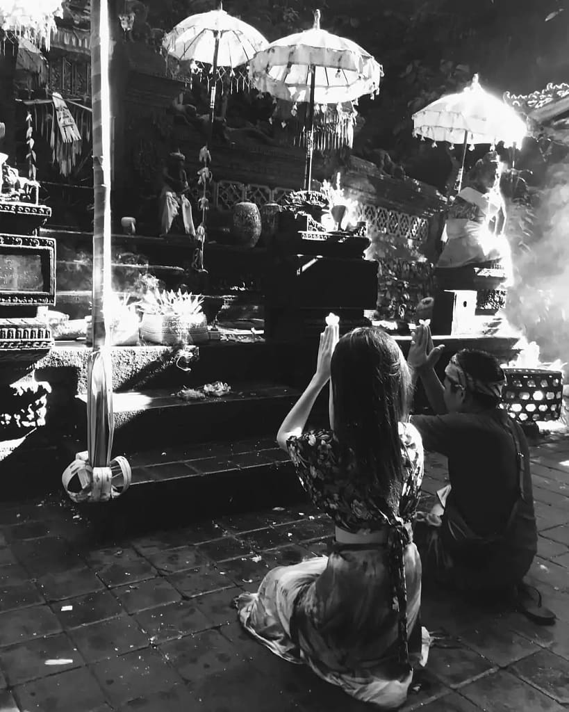 black and white photo of a foreign tourist (female) cross-legged and worshipping (hands on head) in front of a sacred temple accompanied by a pemangku.