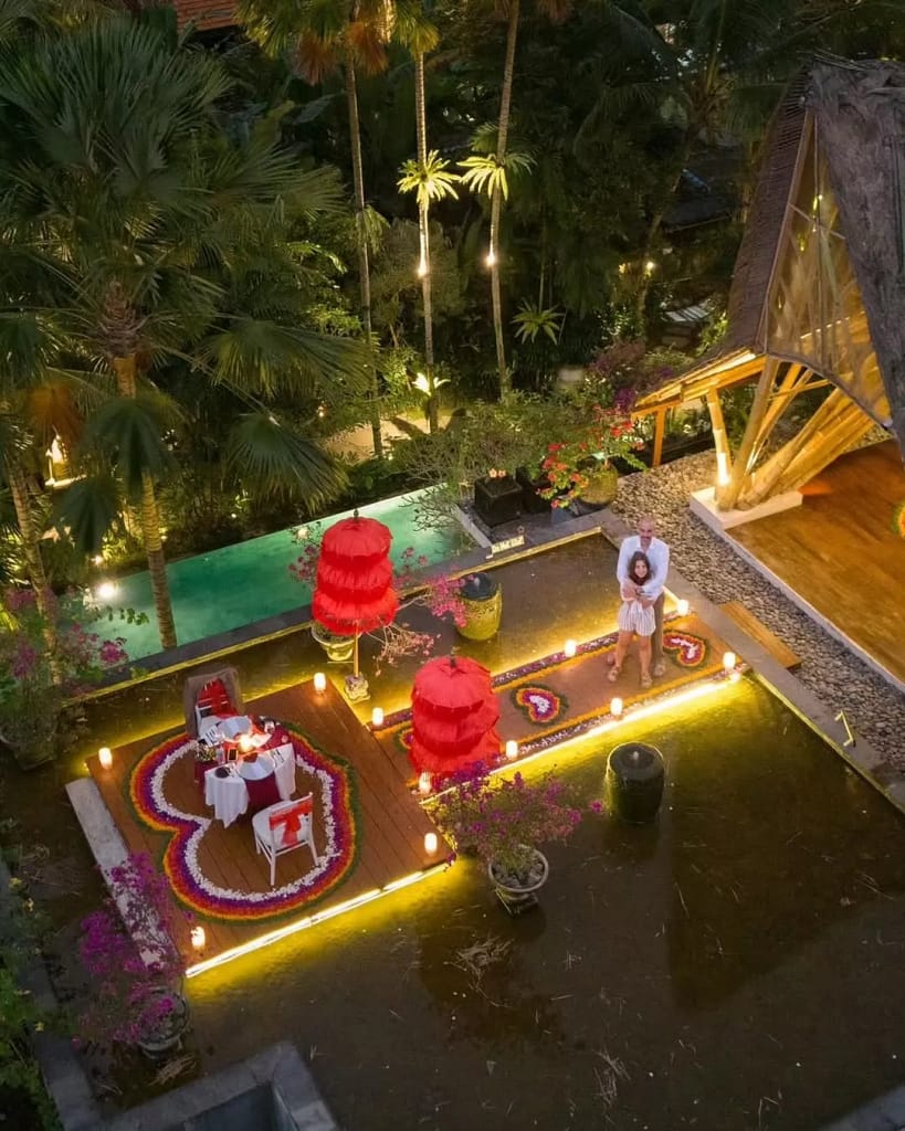 A high-angle view of a luxurious, romantic private dinner setup at a resort or villa in Ubud at night. A couple is standing on a wooden deck, which is decorated with flower petals forming a heart shape and surrounded by numerous lit candles. A dining table for two, set with red accents, is ready for the private, candlelit dinner, with a swimming pool and lush tropical foliage surrounding the area. Why Tourists Love Ubud Bali