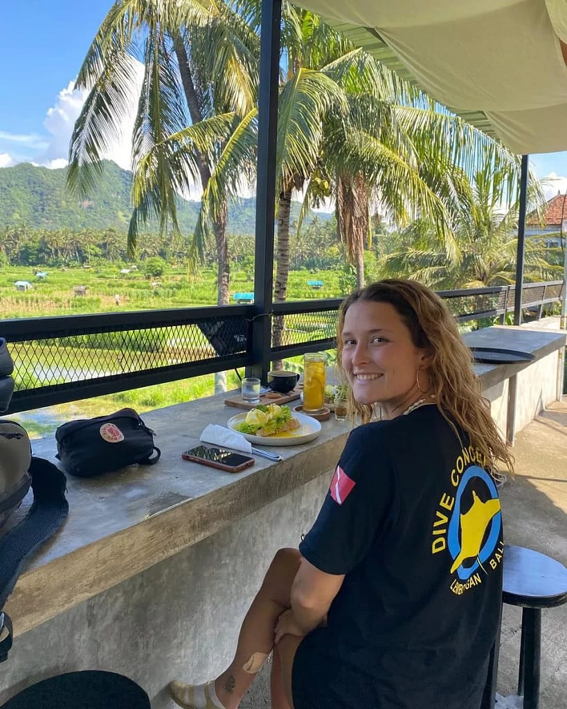 A young woman with long, wavy brown hair, wearing a black t-shirt with a "DIVE CONCEPT BALI" logo on the back, smiling and looking over her shoulder at the camera. She is seated at an outdoor counter enjoying a meal and a drink, with a scenic view of lush green rice fields, palm trees, and distant mountains under a blue sky. Is Ubud Worth Visiting?