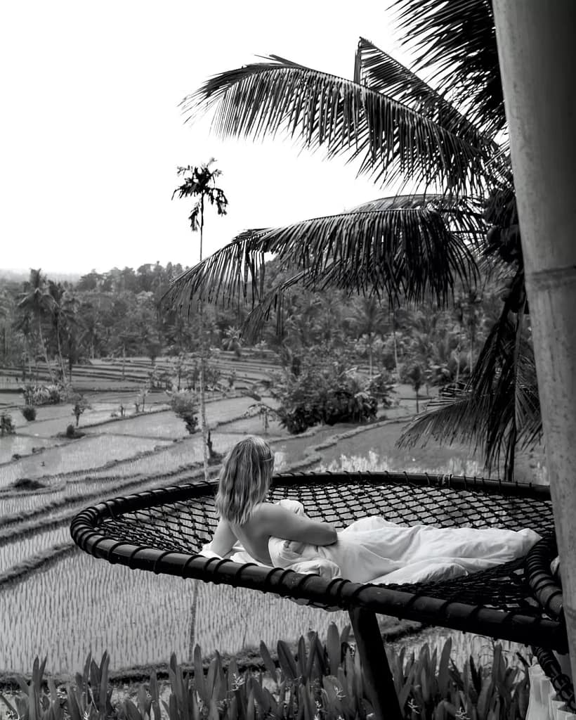 A solitary figure in a white dress relaxes on a net over an extensive landscape of **rice paddies** in this monochrome photograph. The image evokes the sense of **peace and quiet reflection** found when slowing down. Alt: To fall in love with Bali again, consciously uncouple from the algorithm. This quiet moment in the rice fields shows how the answer to is Bali overrated lies in seeking imperfect, unforgettable moments, not perfect photos.