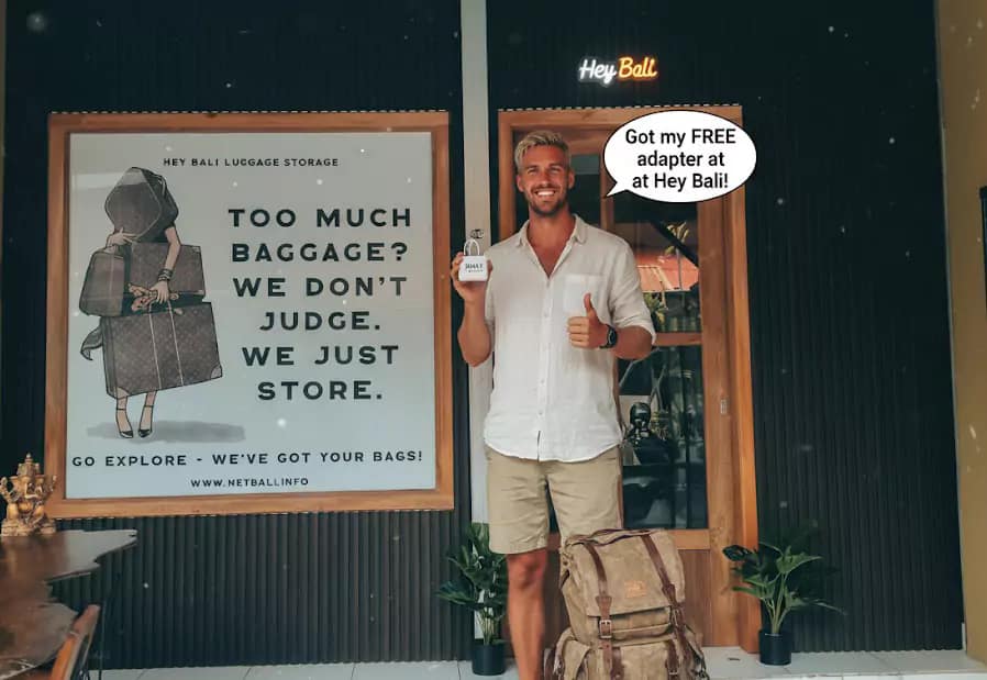 A blonde male traveler from Australia smiles contentedly in front of 'Hey Bali Luggage Storage', holding his newly acquired Free Bali adapter. His travel backpack lies to the side, showing how convenient it is to prepare for a trip to Bali.