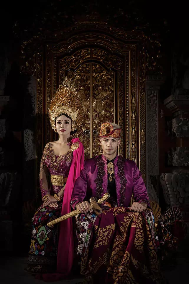 Photo of Balinese groom and bride wearing traditional Balinese wedding clothes sitting like the Balinese king and queen with Balinese carvings in the background.