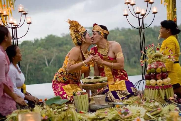 photo of a bule and local couple wearing Balinese clothes kissing after the blessing procession and seen in front of him a lot of banten and offerings in a 5-star hotel in Bali