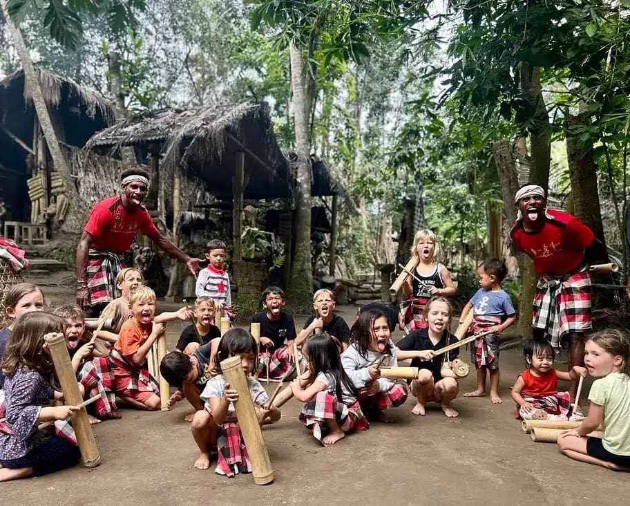A group of diverse children joyfully participating in a traditional Balinese music or play activity outdoors, led by two local men in sarongs. Their genuine smiles and engagement directly challenge the notion that Bali is boring, showcasing the island's vibrant, authentic connections.